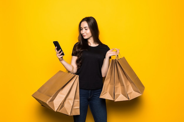 Free Photo beautiful young woman with shopping bags using her smart phone on yellow wall. shopaholic shopping fashion.