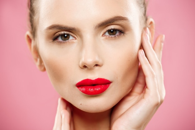 Free photo beauty concept - close up gorgeous young brunette woman face portrait. beauty model girl with bright eyebrows, perfect make-up, red lips, touching her face. isolated on pink background