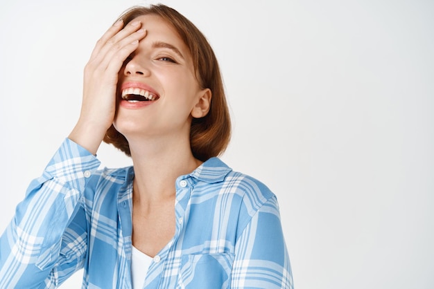 Beauty face Portrait of happy young woman with short hair and natural makeup laughing smiling white teeth covering half of face with hand standing against studio background