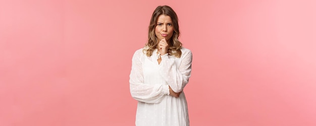 Beauty fashion and women concept Portrait of skeptical and judgemental seriouslooking blond female in white spring dress look disbelief grimacing and frowning pink background