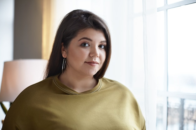 Free photo beauty, femininity, style and fashion concept. portrait of beautiful overweight young dark haired european female with neat make up posing in stylish hotel room interior by window and smiling