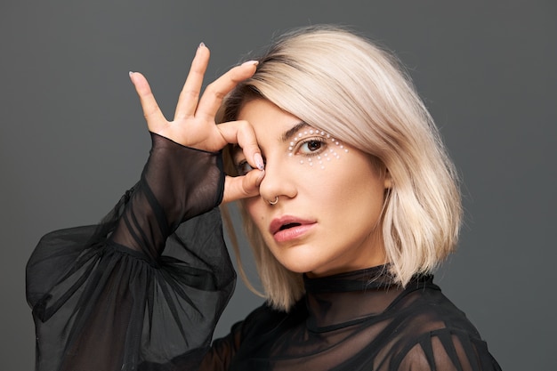 Free photo beauty, glamour, luxury and fashion concept. profile shot of attractive cool young female in stylish transparent black blouse with flares posing isolated, connecting thumb and index finger in ok sign