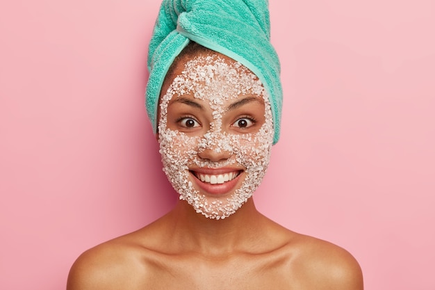 Free photo beauty and health care concept. close up image of satisfied dark skinned female applies white scrub facial mask, cares about purity and softness of her complexion, has turquoise towel on head