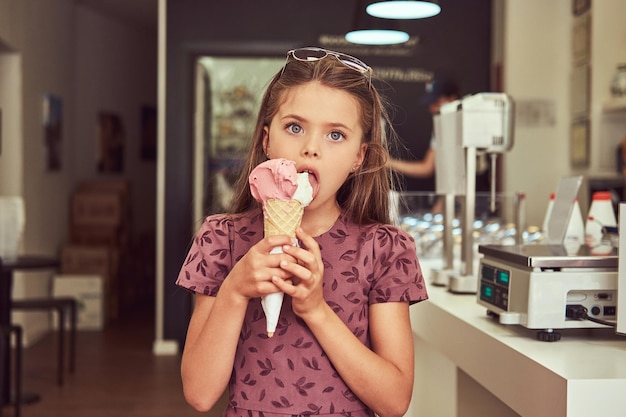 A beauty little girl in a fashionable dress eating strawberry, standing in an ice cream parlor.