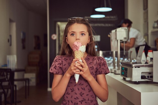 A beauty little girl in a fashionable dress eating strawberry, standing in an ice cream parlor.