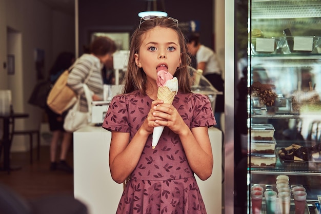 A beauty little girl in a fashionable dress eating strawberry, standing in an ice cream parlor.