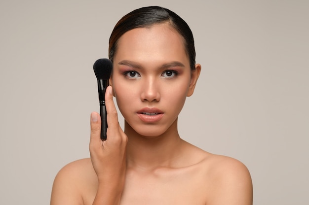 Beauty portrait of asian attractive sensual young woman pose holding makeup blusher brush isolated over gray wall