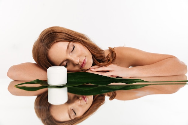 Free photo beauty portrait of ginger woman with long hair lying on mirror table with body cream and green leaf