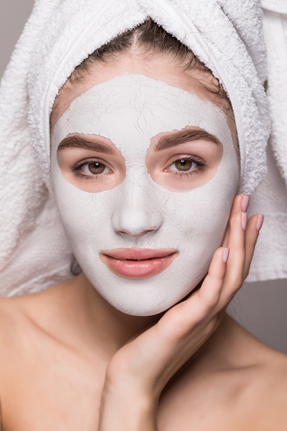 Beauty portrait of woman in towel on head with white nourishing mask or creme on face, white wall isolated. Skincare cleansing eco organic cosmetic spa relax concept