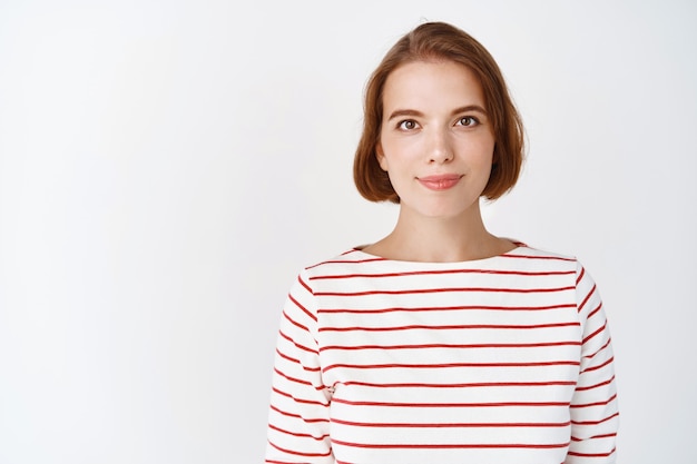 Beauty. Portrait of young confident woman with natural light facial skin, smile tenderly , standing in striped blouse against white wall