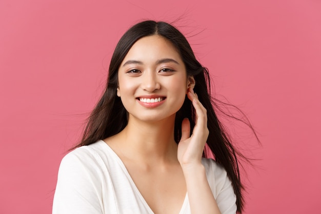 Beauty salon, haircare and skincare products advertisement concept. Close-up of beautiful young asian woman smiling as wind softly blowing at haircut, standing pink background.