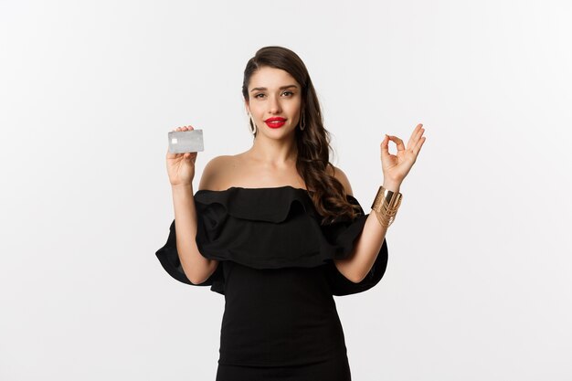 Beauty and shopping concept. Gorgeous woman in luxury jewelry and black dress, showing okay sign and credit card, standing over white background.