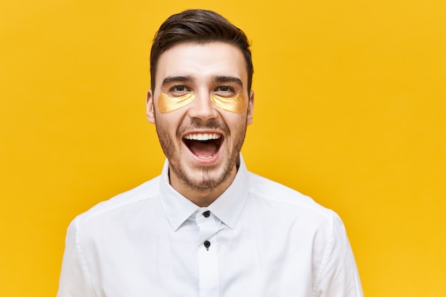 Beauty and skin care concept. Picture of excited joyful young male with stubble wearing golden patches under eyes to look awake and refreshed, screaming with excitement, opening mouth wide opened