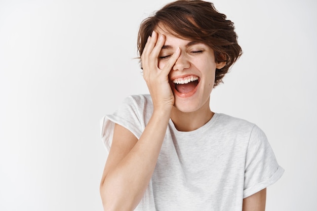 Beauty and skincare. Portrait of happy caucasian woman with short hair, touching clean smooth facial skin and laughing carefree, standing with eyes closed on white wall