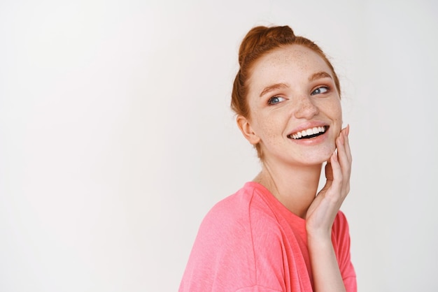 Beauty and skincare Smiling girl with ginger hair combed in messy bun touching perfect and glowing skin and smiling showing face without makeup white background