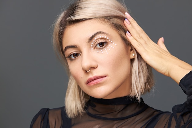 Beauty, style and femininity concept. gorgeous sensual European female with blonde bob hairstyle demonstrating her stylish make up, white crystals around eye