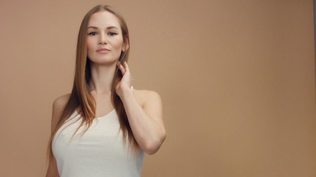 Beauty woman model in studio on beige background touches her straight blonde hair and watching at camera