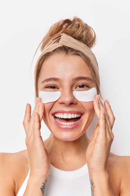 Beauty woman with combed hair applies patches under eyes for perfect skin has refreshed well cared complexion smiles broadly wears headband undergoes facial treatment isolated over white background