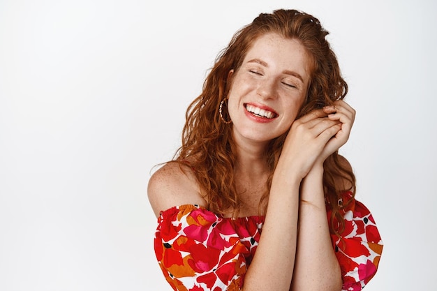 Free photo beauty and women concept dreamy smiling woman with red curly hairstyle close eyes and grinning carefree dreaming of something standing in dress against white background