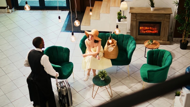 Free Photo bellhop offering services to woman in lounge area, waiting to do room check in at seaside hotel. tourist sitting on couch and talking to hotel concierge, summer clothes. handheld shot.