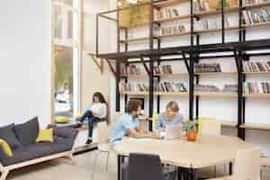 Free photo big modern library in morning. two people sitting, looking in laptop monitor talking about startup project. girl sitting on window sill reading book on smartphone spending time before study.