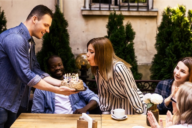 Free photo birthday cake with burning candles which girl is blowing out and best friends at the terrace of a cosy cafe