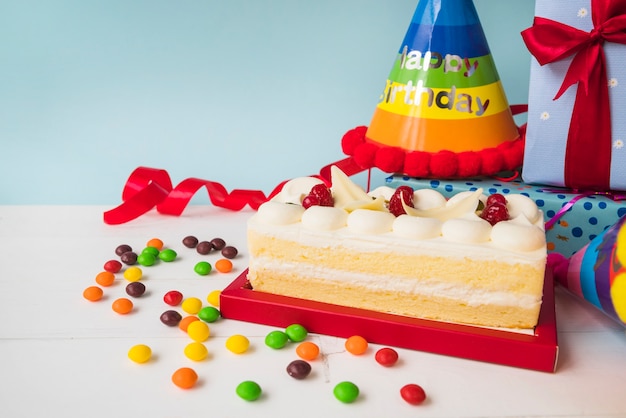 Free photo birthday cake with candies; hat; and presents on table against blue backdrop