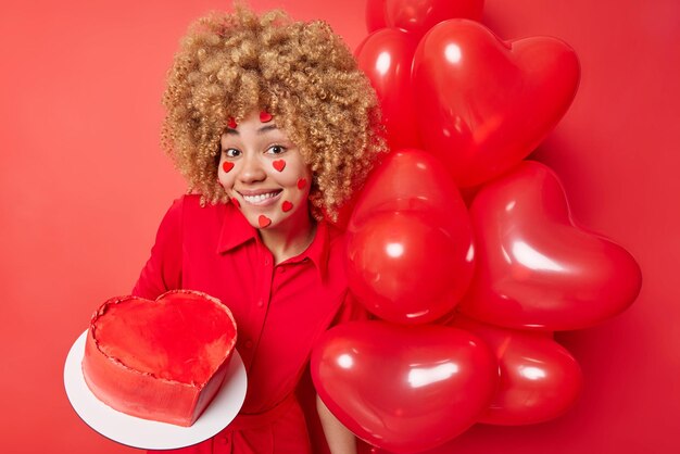 Birthday celebration concept Smiling European woman with curly blonde hair wears festive dress being in good mood holds cake and bunch of inflated balloons isolated over vivid red background