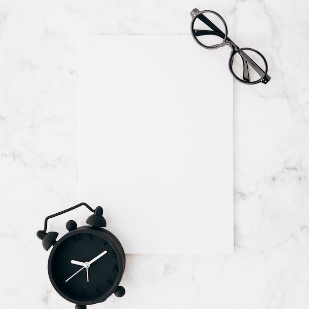 Free photo black alarm clock and eyeglasses on white blank paper against marble textured background