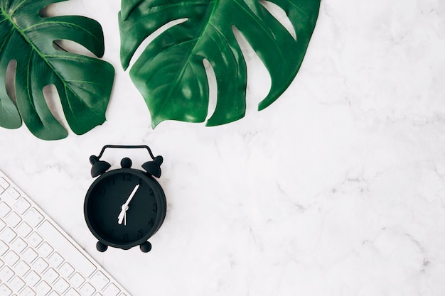 Free photo black alarm clock; keyboard and green monstera leaves on white marble background