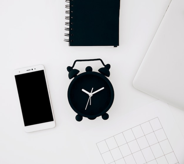 Free photo black alarm clock; spiral notepad; smartphone; page and laptop on white desk