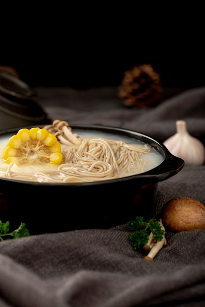 Free photo black bowl filled with noodle soup and corn on a grey cloth with garlic and mushrooms