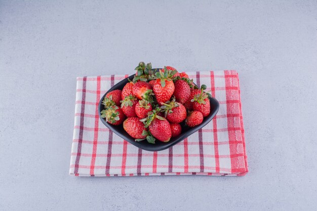 Black bowl with a heap of strawberries on marble background.