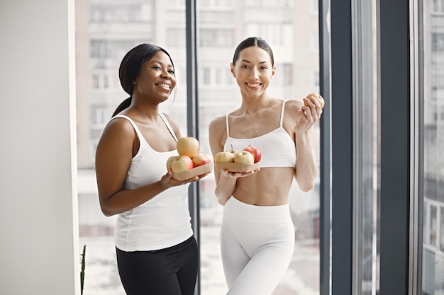 Black and caucasian women standing in studio with big windows and holding apples