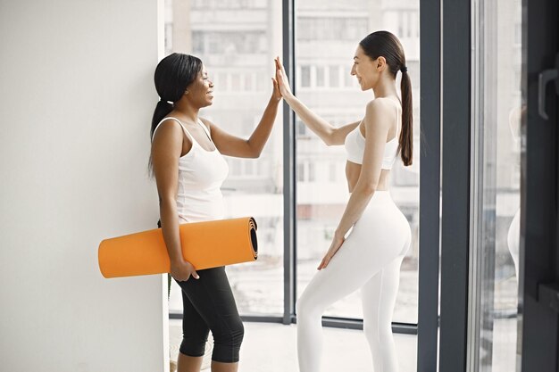 Black and caucasian women standing with rolling mat in studio with big windows