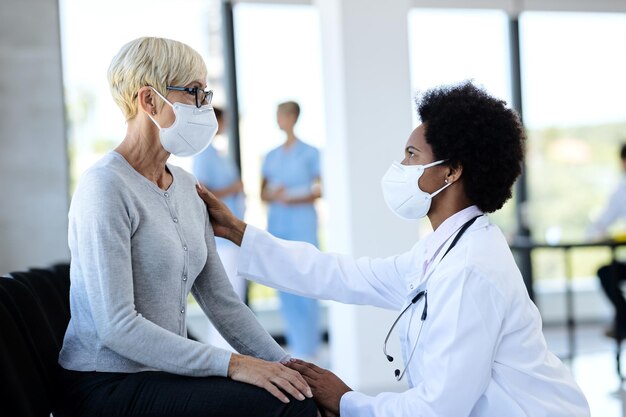 Black doctor and mature patient wearing protective face masks while talking in waiting room at clinic