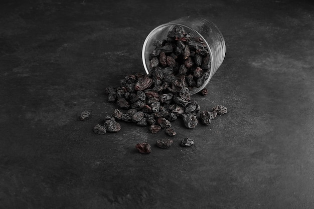 Black dry sultanas out of a glass cup on black surface. 