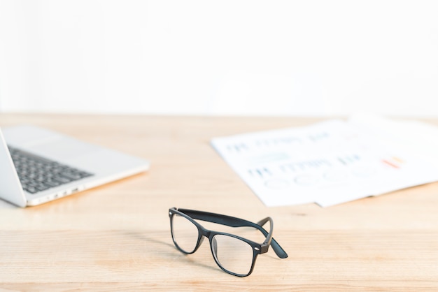 Black eyeglasses in front of laptop and graph on wooden desk