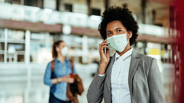 Black female passenger talking on smart phone while wearing face mask at the airport during virus epidemic