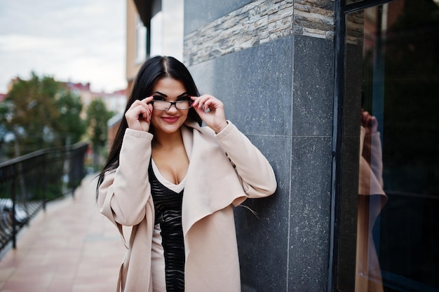 Black hair sexy woman in glasses and coat posed against building with modern windows