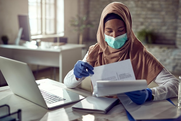 Black Islamic businesswoman wearing protective face mask while going through paperwork in the office during virus pandemic