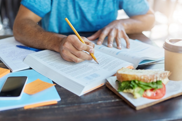 Free photo black male student underlining important information in textbook using pencil while making history research at university canteen during lunch; phone, coffee and food resting on table