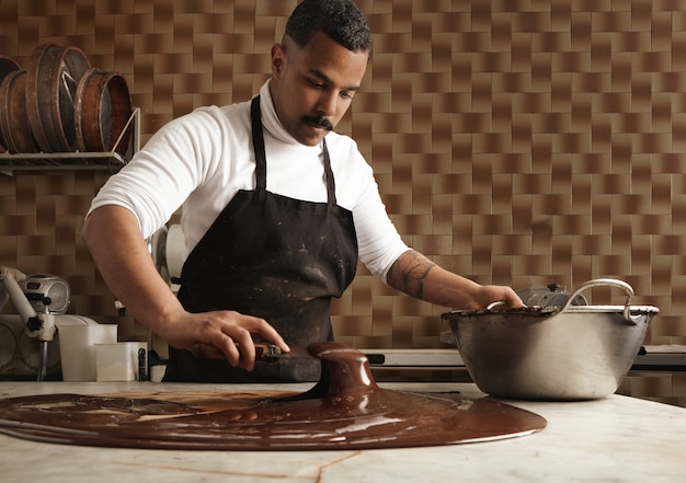 Free Photo black man professional chief collects tasty melted chocolate from vintage marble table to big steel pot in his artisan rustic kitchen with industrial retro machines.