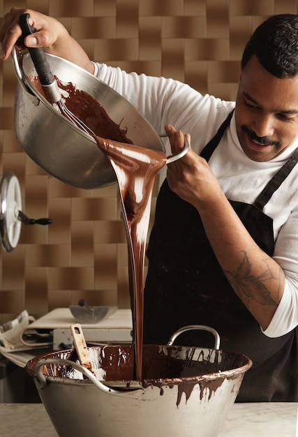 Free photo black man professional chief pours tasty melted chocolate from one big steel pot to another before making chocolate bars