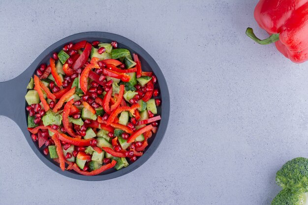 Black pan of vegetable salad next to a bell pepper and a broccoli on marble background. High quality photo