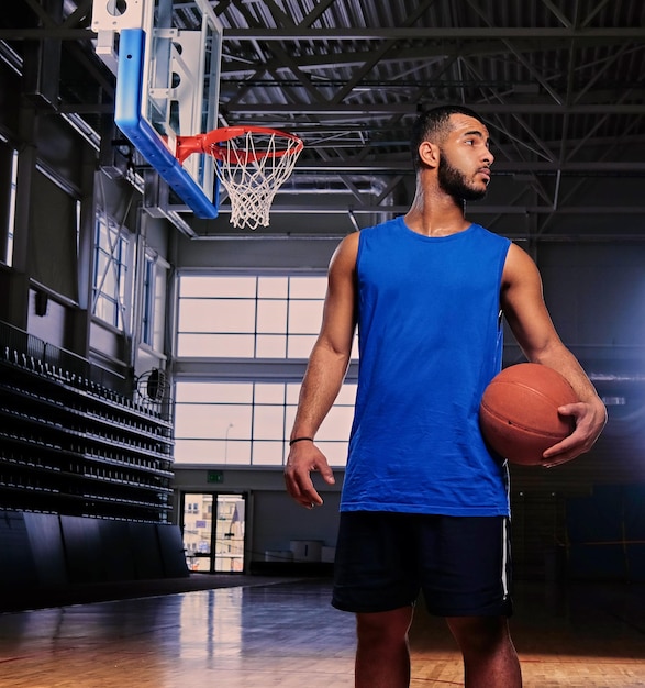 Black professional basketball player holds a ball over the hoop in a game hall.