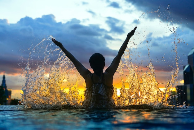 Black silhouette of asian woman splash water on summer vacation holiday relaxing in infinity swimming pool with blue sea sunset view with high rise skyscape urban downtown Healthy happiness lifestyle