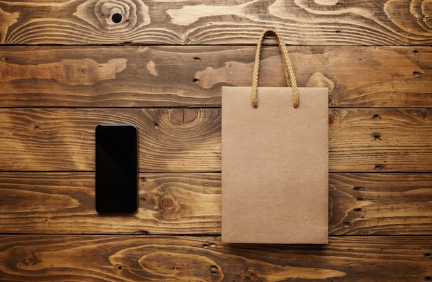 black smarthpone lying next to a craft paper bag with light brown string handles on a beautiful wooden table, shot from top