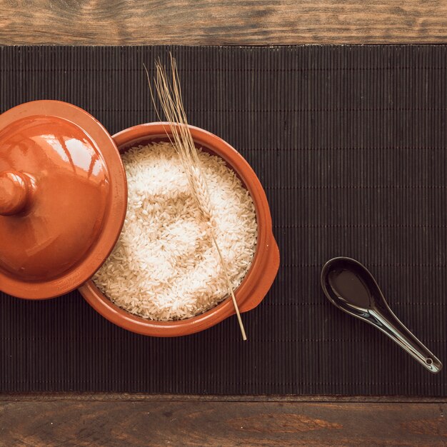 Black spoon with uncooked rice grains pot with lid on place mat over wooden table