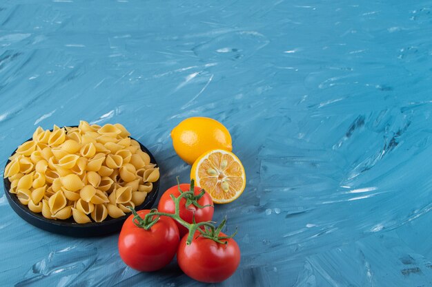 A black wooden plate of raw pasta with lemon and fresh red tomatoes. 